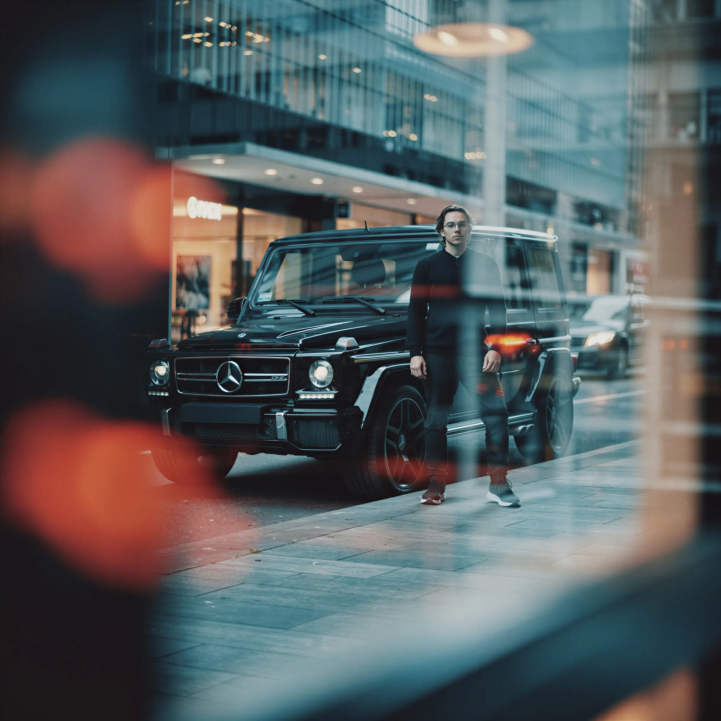 A man beside a black G-Class on a city street, seen through reflections and soft light