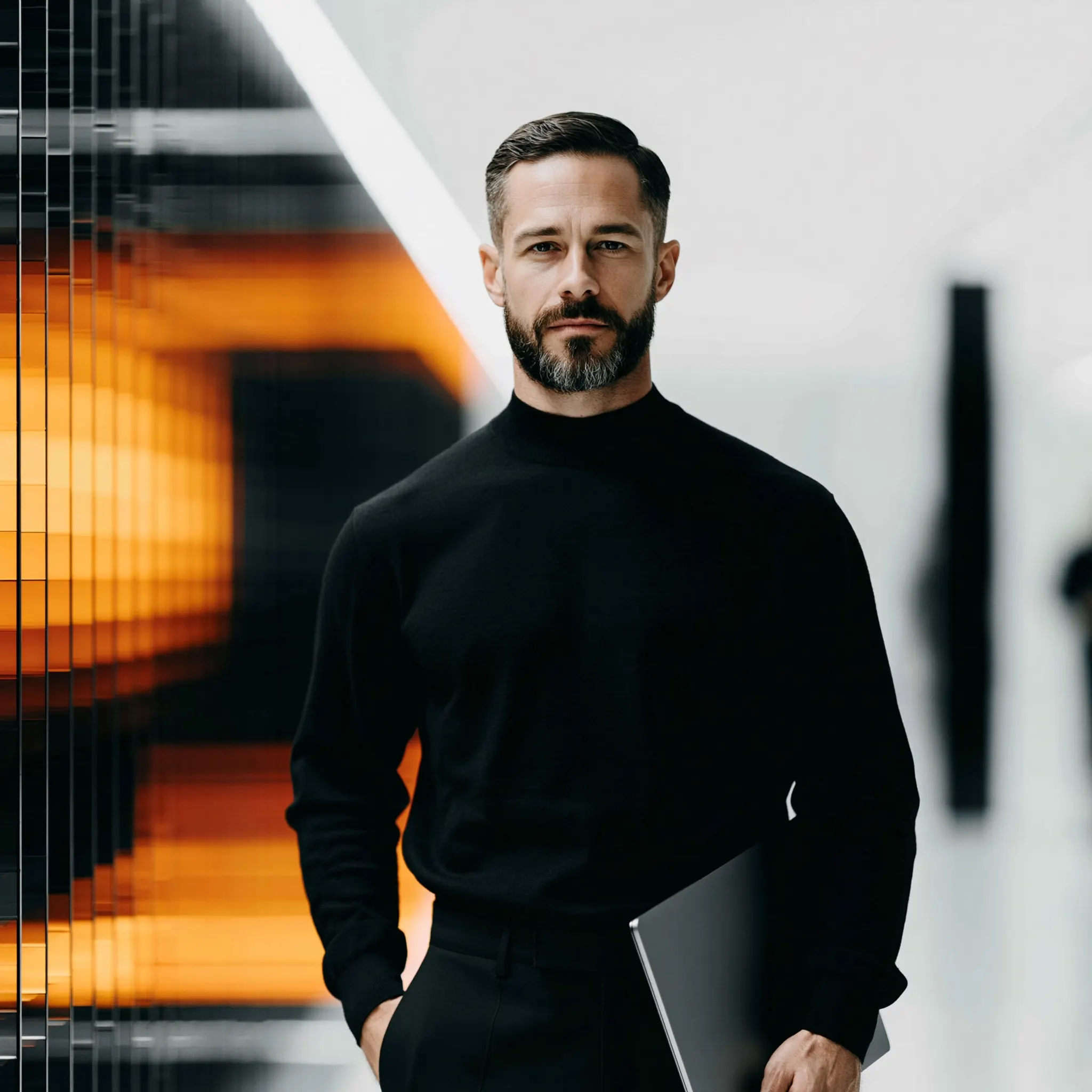 A man in a black roll-neck holding a laptop, with blurred orange light behind him